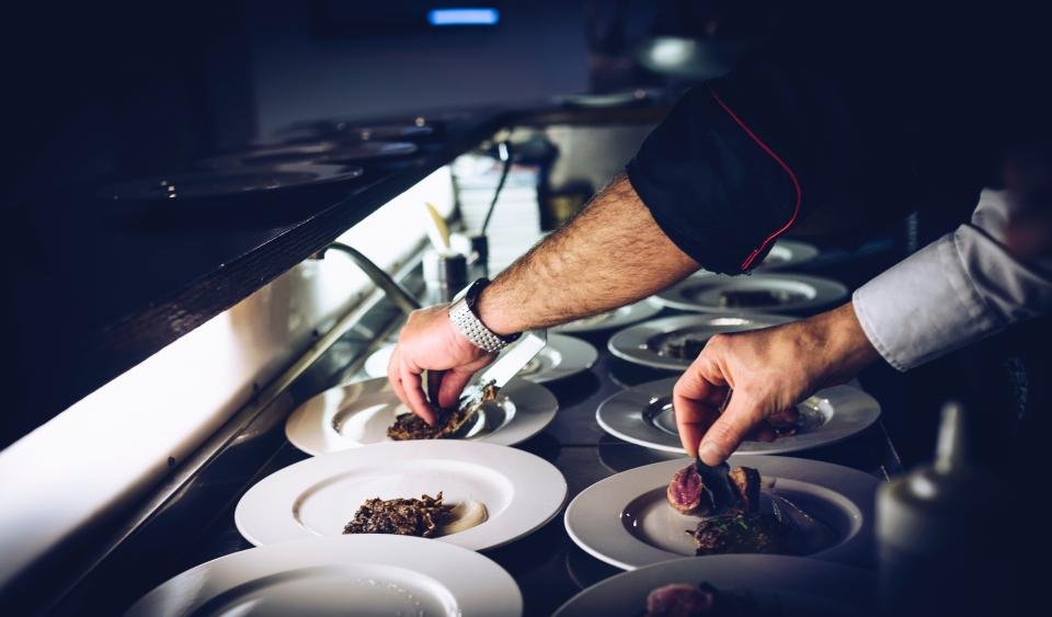 Manos de cocina emplatando varios platos a la vez, en el pase bajo las lámparas de calor.