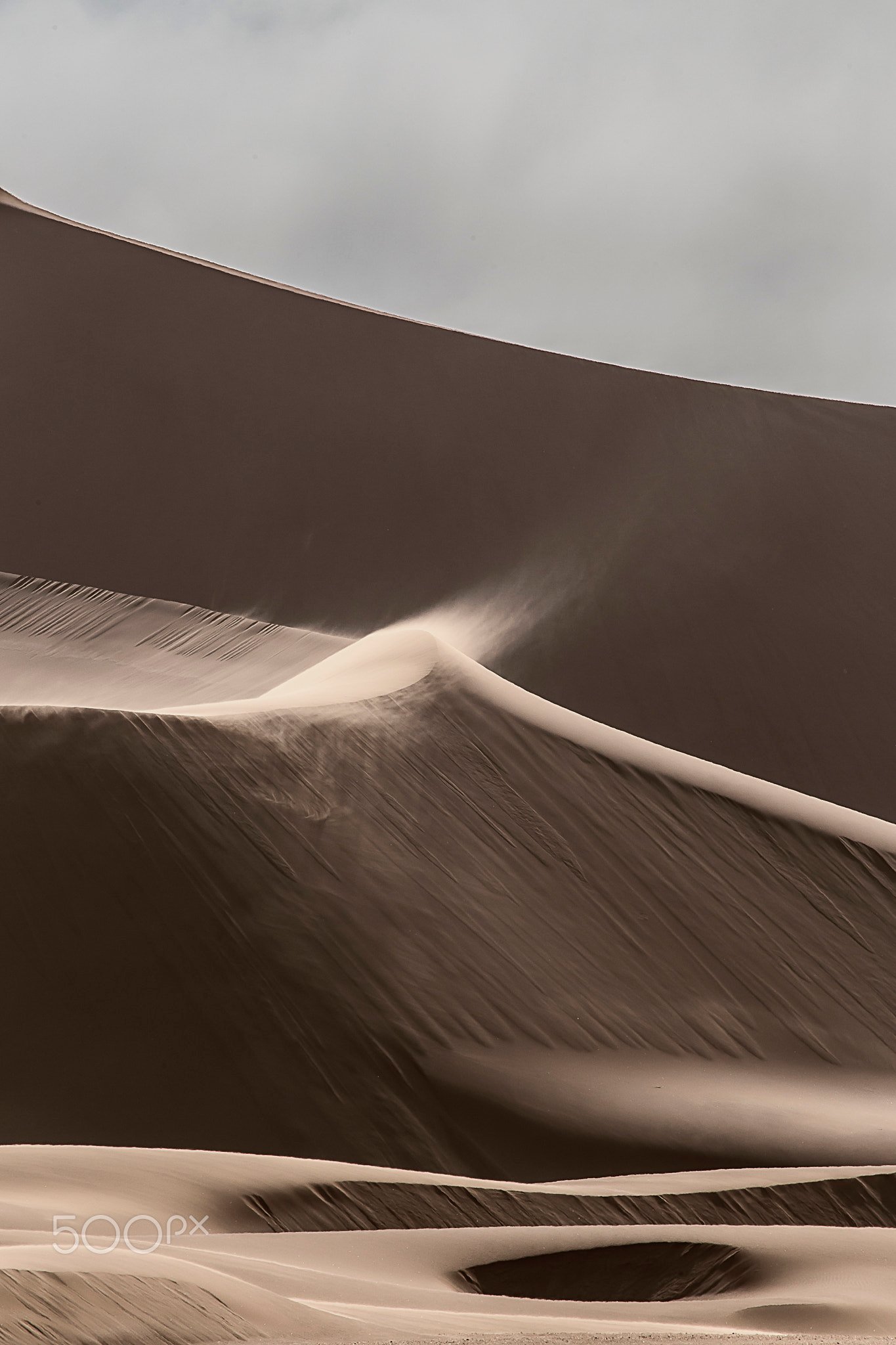 Duna de arena con viento levantando el polvo de la cresta, tonos tierra