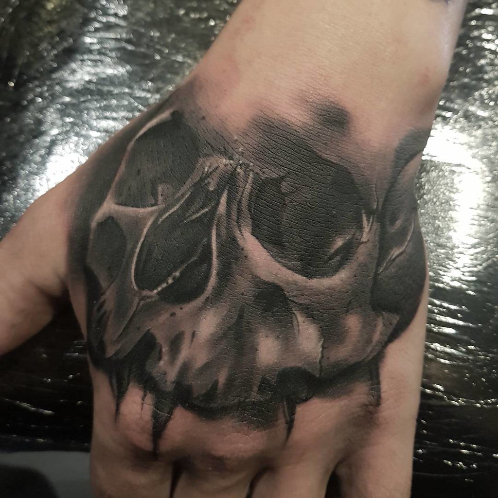 Cráneo hiperrealista tatuado en el dorso de una mano, negro y gris con textura de hueso